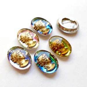 K&ouml;nnte beinhalten: Sechs ovale Glascabochons mit schillernden Regenbogenfarben. Jeder Cabochon zeigt ein goldfarbenes Profil eines menschlichen Gesichts.