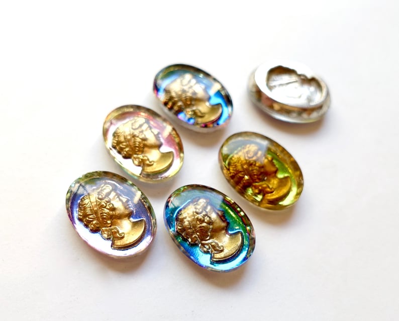 K&ouml;nnte beinhalten: Sechs ovale Glascabochons mit schillernden Regenbogenfarben. Jeder Cabochon zeigt ein goldfarbenes Profil eines menschlichen Gesichts.