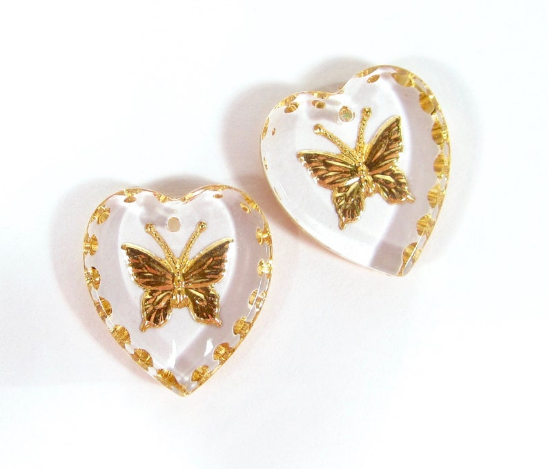 Puede incluir: Dos colgantes transparentes en forma de coraz&oacute;n con detalles de mariposas doradas. Las mariposas est&aacute;n centradas dentro de los corazones, y los bordes est&aacute;n adornados con puntos dorados. Cada colgante tiene un peque&ntilde;o agujero en la parte superior para colgar.