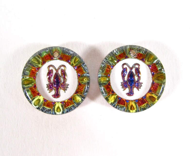 May include: A pair of round, iridescent earrings with a lobster design in the center. The earrings have a multi-colored, mosaic-like pattern around the edge.