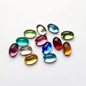 Könnte beinhalten: Sortiment ovaler Glascabochons in verschiedenen Farben, darunter rosa, blau, grün, rot und gelb. Die Cabochons sind auf einer weißen Oberfläche angeordnet.