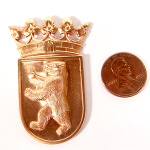 Könnte beinhalten: Ein goldfarbener Metallanhänger mit dem Berliner Stadtwappen. Der Anhänger zeigt einen Bären, der auf seinen Hinterbeinen steht, mit einer Krone über dem Bären. Der Anhänger ist etwa so groß wie ein deutscher Cent.