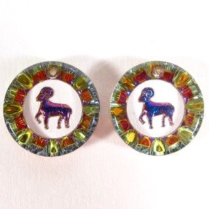 May include: A pair of iridescent glass earrings with a gold trim and a white background. Each earring features a stylized ram, the astrological symbol for Aries.