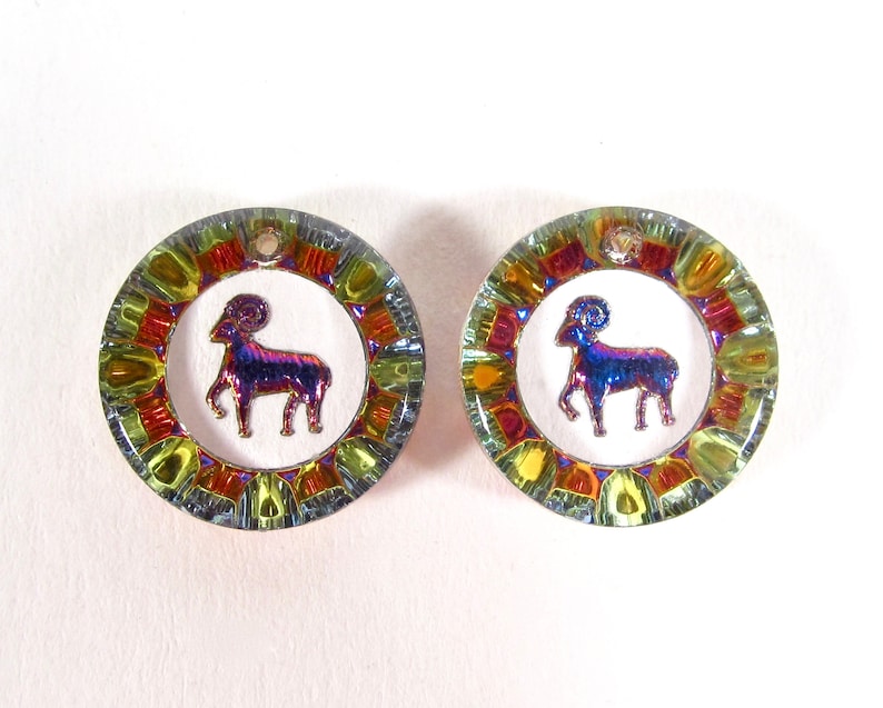May include: A pair of iridescent glass earrings with a gold trim and a white background. Each earring features a stylized ram, the astrological symbol for Aries.