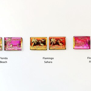 May include: Rectangular, iridescent Florida-themed magnets. Designs include "Florida Beach" with flamingos, "Flamingo Sahara" with palm trees and flamingos, and "Florida Flag" with text. Each magnet has a gold-colored border.