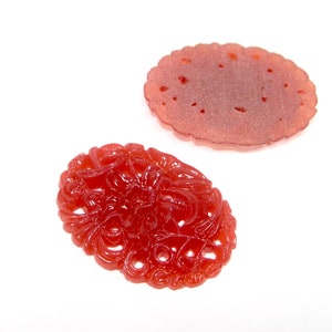 May include: Two red carved jade pendants. One pendant is a simple oval shape with a smooth surface. The other pendant is a more intricate design with a floral pattern.