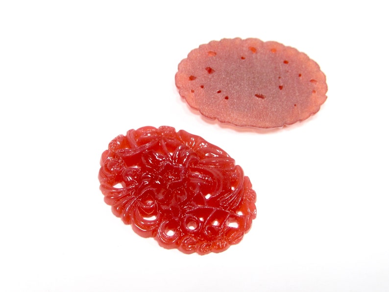 May include: Two red carved jade pendants. One pendant is a simple oval shape with a smooth surface. The other pendant is a more intricate design with a floral pattern.