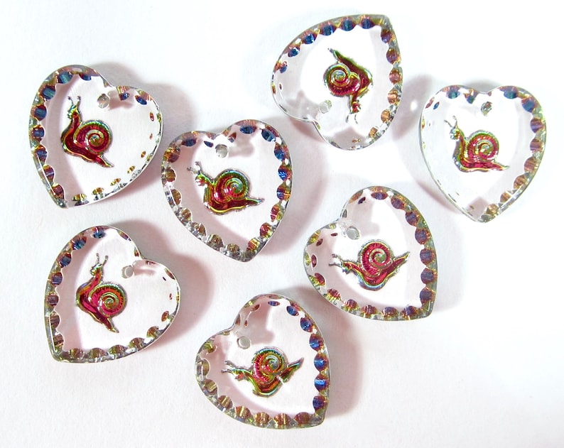 May include: Seven iridescent heart-shaped buttons with a snail design. The buttons are clear with a silver trim and a white background. Each button has a small hole at the top for sewing.