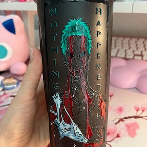Puede incluir: Vaso negro con una ilustraci&oacute;n detallada de un personaje con cabello verde y el texto "NOTHING HAPPENED" en verde. La obra de arte incluye detalles en rojo y blanco, creando un contraste visual llamativo.