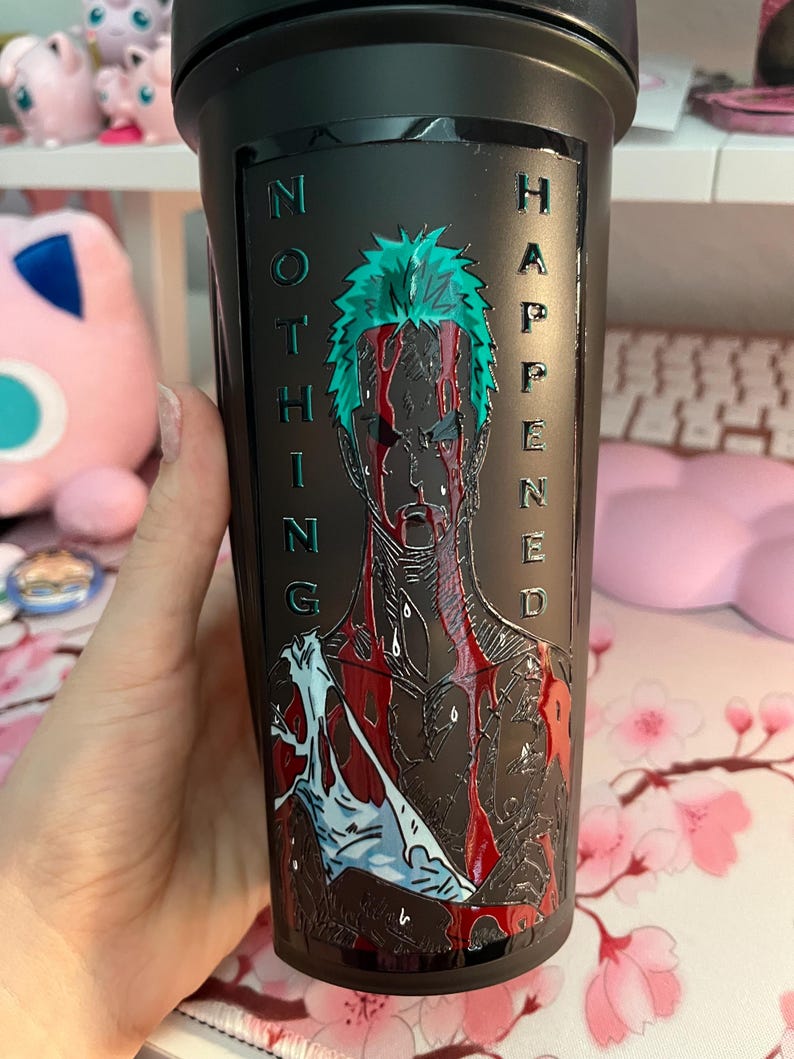 Puede incluir: Vaso negro con una ilustraci&oacute;n detallada de un personaje con cabello verde y el texto "NOTHING HAPPENED" en verde. La obra de arte incluye detalles en rojo y blanco, creando un contraste visual llamativo.