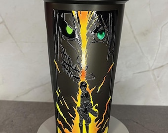 Könnte beinhalten: Schwarze Shaker-Flasche mit einem Grafikdruck eines Charakters aus dem Anime Attack on Titan. Der Charakter hat grüne Augen und ist von gelben und orangefarbenen Flammen umgeben.