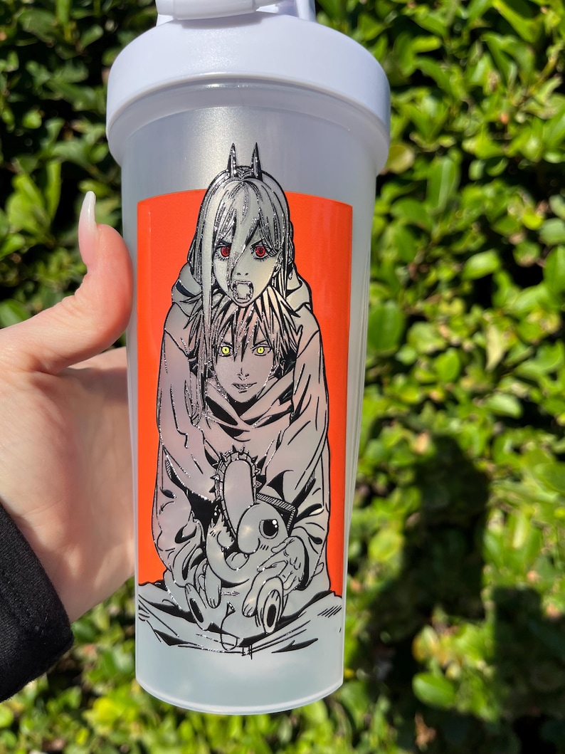Pu&ograve; includere: Una bottiglia shaker in plastica trasparente con coperchio bianco. La bottiglia presenta un adesivo arancione vibrante con un'illustrazione in stile manga di due personaggi. Un personaggio ha gli occhi rossi e l'altro gli occhi gialli. L'illustrazione &egrave; delineata in nero.