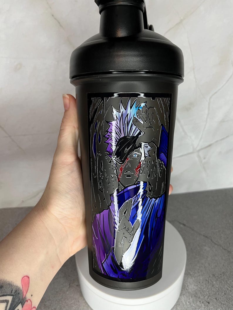 May include: Black shaker bottle with a graphic of a character from the anime Naruto. The character has white hair, blue eyes, and is wearing a purple and blue outfit. The background is black and white.