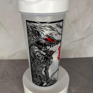 May include: White plastic shaker bottle with a black and white graphic of a monster with red eyes. The bottle has a white lid with a handle. The graphic is on a clear plastic body.