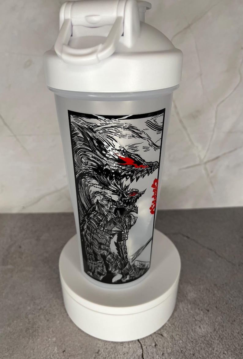 May include: White plastic shaker bottle with a black and white graphic of a monster with red eyes. The bottle has a white lid with a handle. The graphic is on a clear plastic body.