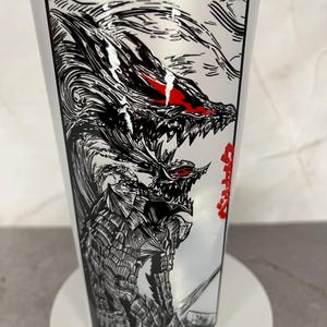 May include: A white plastic tumbler featuring a detailed black and white illustration of a fierce dragon-like creature with red eyes. The design is reminiscent of manga or anime art, with Japanese text on the side.