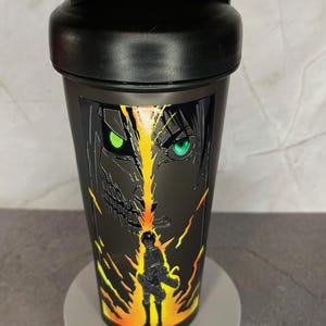 May include: Black shaker bottle with a graphic of a character from the anime Attack on Titan. The character has green eyes and is surrounded by yellow and orange flames.
