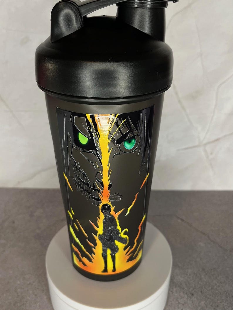 May include: Black shaker bottle with a graphic of a character from the anime Attack on Titan. The character has green eyes and is surrounded by yellow and orange flames.