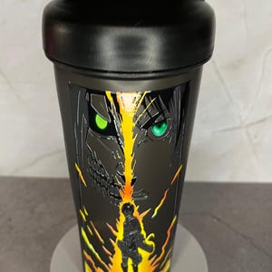May include: Black shaker bottle with a graphic of a character from the anime Attack on Titan. The character has green eyes and is surrounded by flames.
