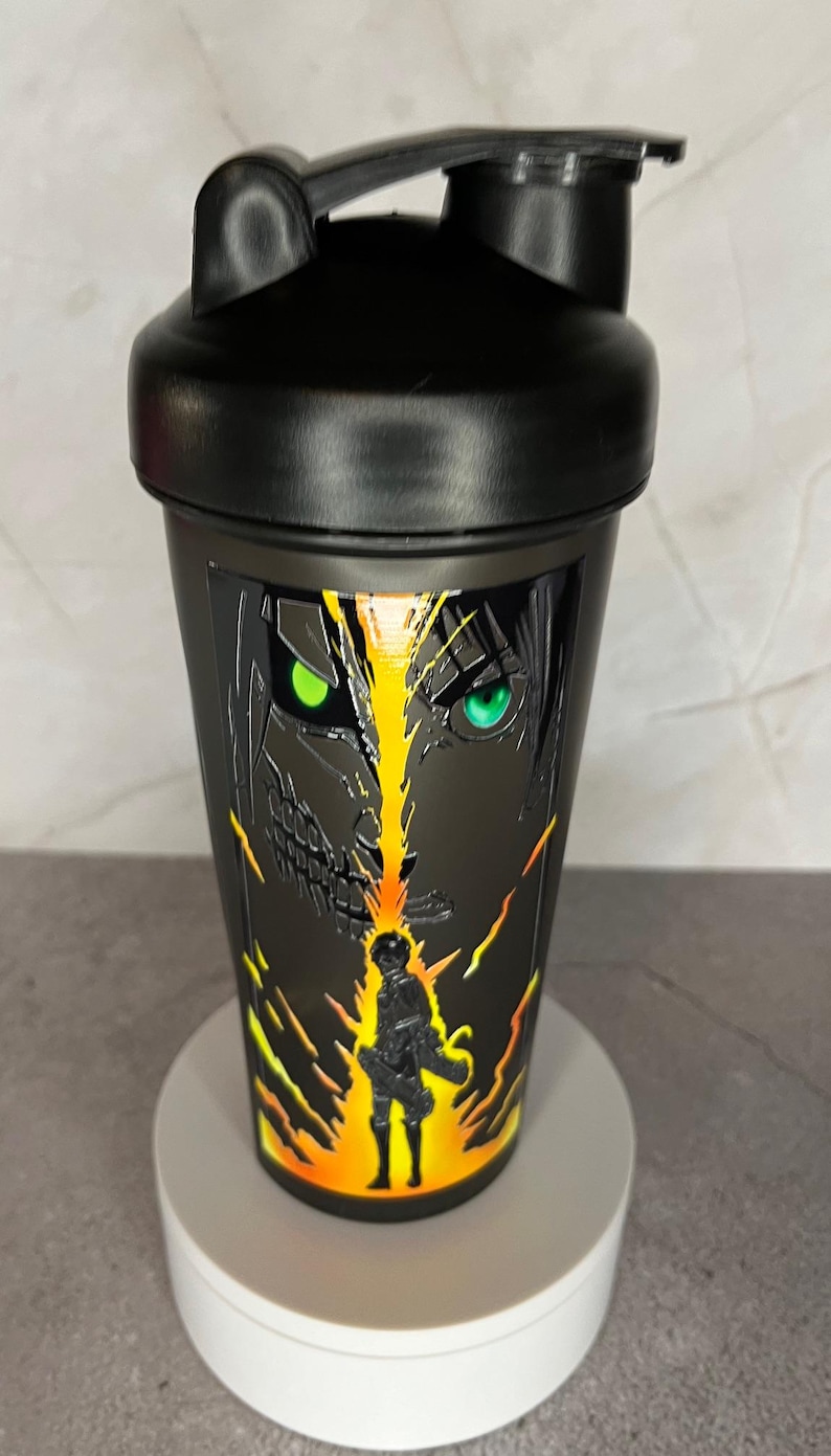 May include: Black shaker bottle with a graphic of a character from the anime Attack on Titan. The character has green eyes and is surrounded by flames.