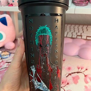 Puede incluir: Botella agitadora negra con tapa de rosca. La botella presenta un gr&aacute;fico de un personaje con cabello verde y marcas rojas similares a sangre, con las palabras "NOTHING HAPPENED" en relieve en el lateral.
