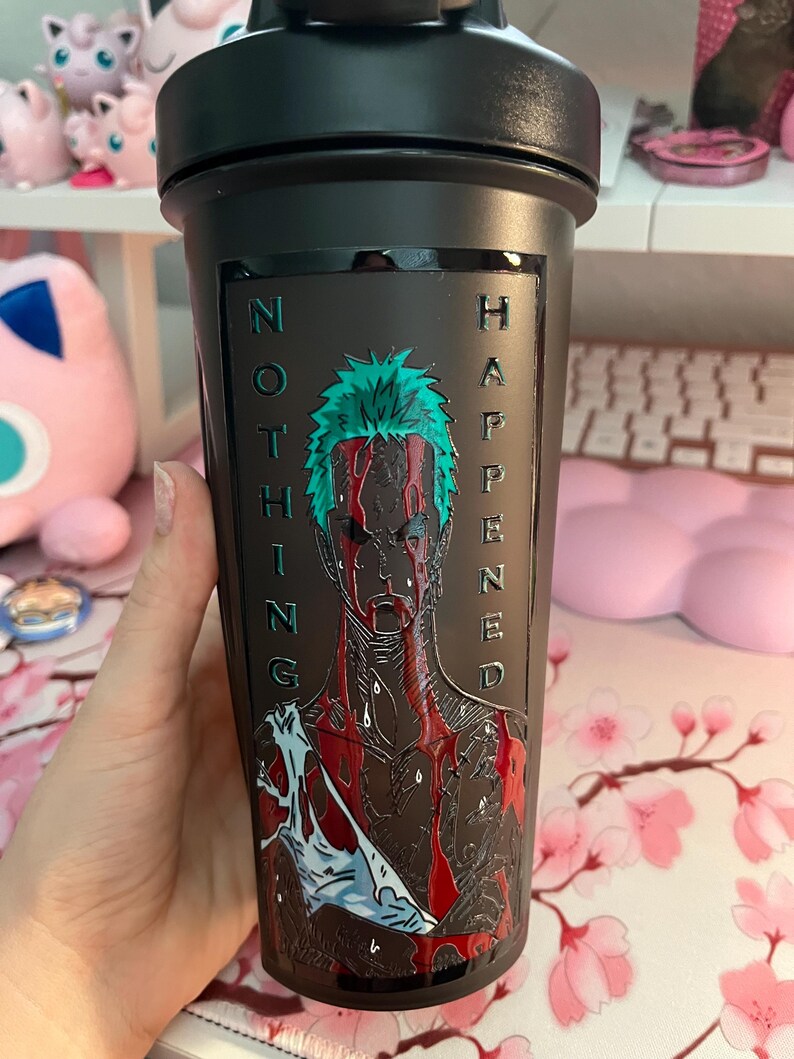 Puede incluir: Botella agitadora negra con tapa de rosca. La botella presenta un gr&aacute;fico de un personaje con cabello verde y marcas rojas similares a sangre, con las palabras "NOTHING HAPPENED" en relieve en el lateral.