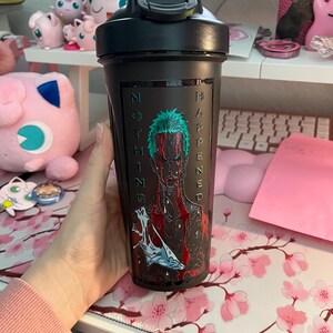 Puede incluir: Botella agitadora negra con tapa abatible. La botella presenta un gr&aacute;fico de un personaje con cabello verde y las palabras "NOTHING HAPPENED" en blanco. La botella se sostiene frente a un escritorio con un mousepad floral.