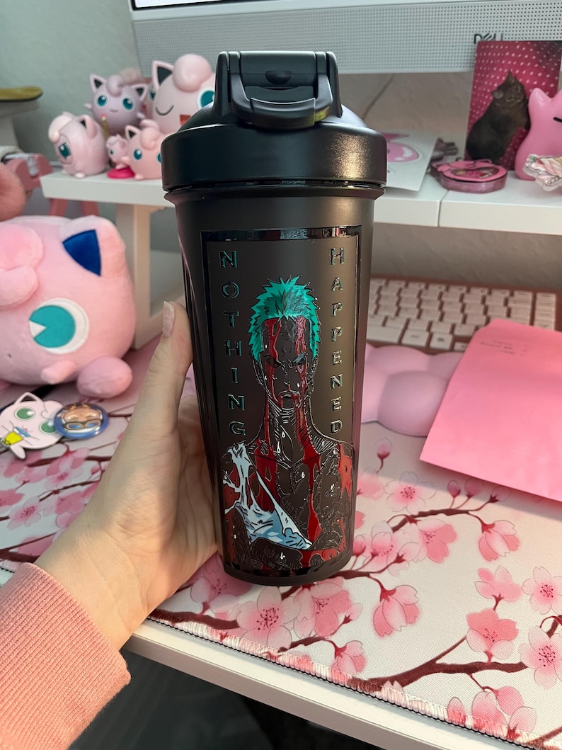 Puede incluir: Botella agitadora negra con tapa abatible. La botella presenta un gr&aacute;fico de un personaje con cabello verde y las palabras "NOTHING HAPPENED" en blanco. La botella se sostiene frente a un escritorio con un mousepad floral.