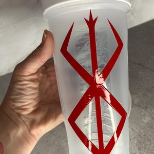 May include: A white and clear plastic shaker bottle with a red graphic design. The bottle has a white lid and a red symbol on the front. The shaker is held by a hand.