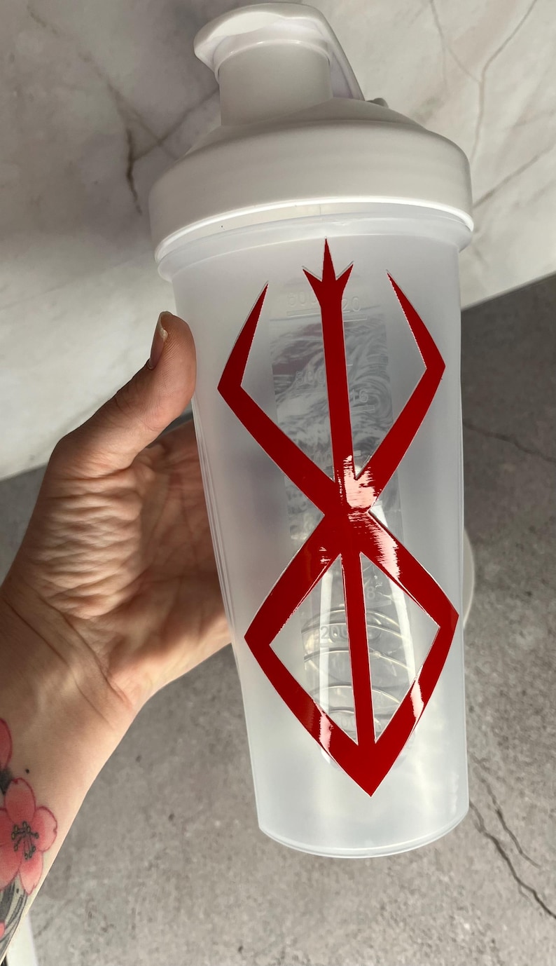May include: A white and clear plastic shaker bottle with a red graphic design. The bottle has a white lid and a red symbol on the front. The shaker is held by a hand.