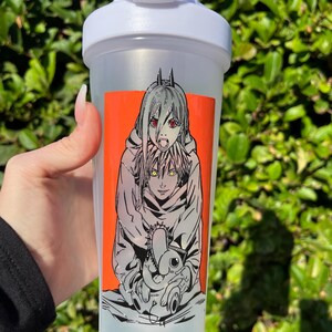 Pu&ograve; includere: Una bottiglia shaker in plastica trasparente con coperchio bianco. La bottiglia presenta una vivace grafica rettangolare arancione con un'illustrazione in stile manga di due personaggi. Uno ha i capelli argentati e gli occhi rossi.