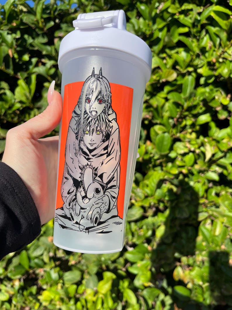 Pu&ograve; includere: Una bottiglia shaker in plastica trasparente con coperchio bianco e chiusura a ribalta. La bottiglia presenta una vivace grafica rettangolare arancione con un'illustrazione in bianco e nero in stile anime di due personaggi. Lo sfondo &egrave; verde sfocato.