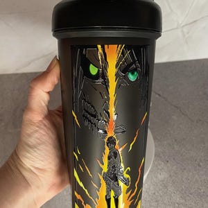 May include: Black shaker bottle with a graphic of a character from the anime Attack on Titan. The character has green eyes and is surrounded by yellow and orange flames.