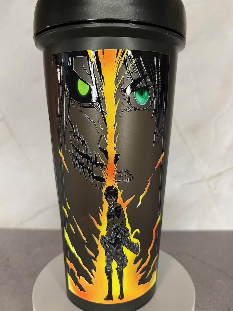 May include: Black shaker bottle with a graphic of a character from the anime Attack on Titan. The character is depicted with a fierce expression and glowing yellow and orange energy behind them.