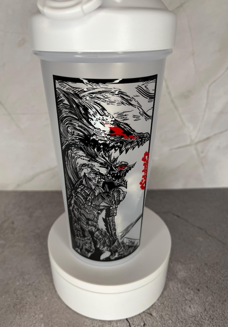 May include: A white and clear plastic shaker bottle featuring a black and white illustration of a monster with red eyes and teeth. The bottle has a white lid and is on a white stand.