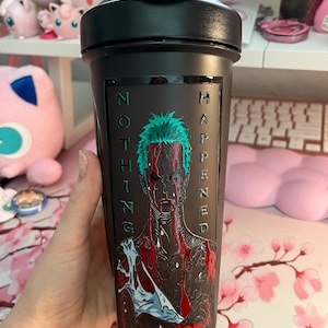 Puede incluir: Botella agitadora negra con tapa abatible. La botella presenta un gr&aacute;fico de un personaje con cabello verde y las palabras "NOTHING HAPPENED" en texto vertical. El personaje est&aacute; representado con marcas rojas similares a la sangre.