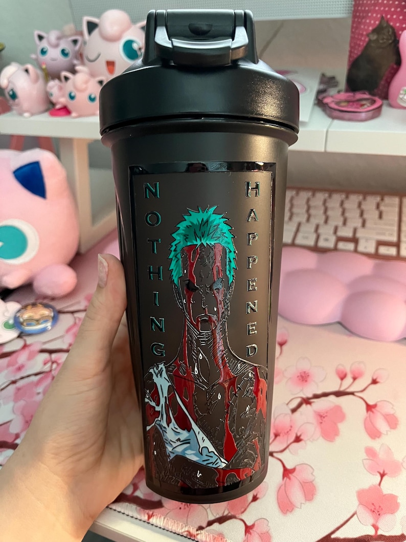 Puede incluir: Botella agitadora negra con tapa abatible. La botella presenta un gr&aacute;fico de un personaje con cabello verde y las palabras "NOTHING HAPPENED" en texto vertical. El personaje est&aacute; representado con marcas rojas similares a la sangre.