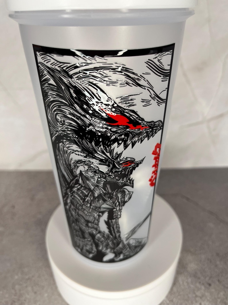 May include: A clear plastic tumbler with a white lid. The tumbler features a black and white illustration of a monster with red eyes and Japanese text. The design wraps around the cup.