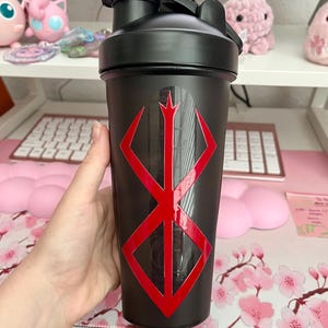 May include: Black shaker bottle with a red geometric design. The bottle has a black lid and a clear measurement scale on the side. The design is a red, stylized symbol. The bottle is held in a hand.