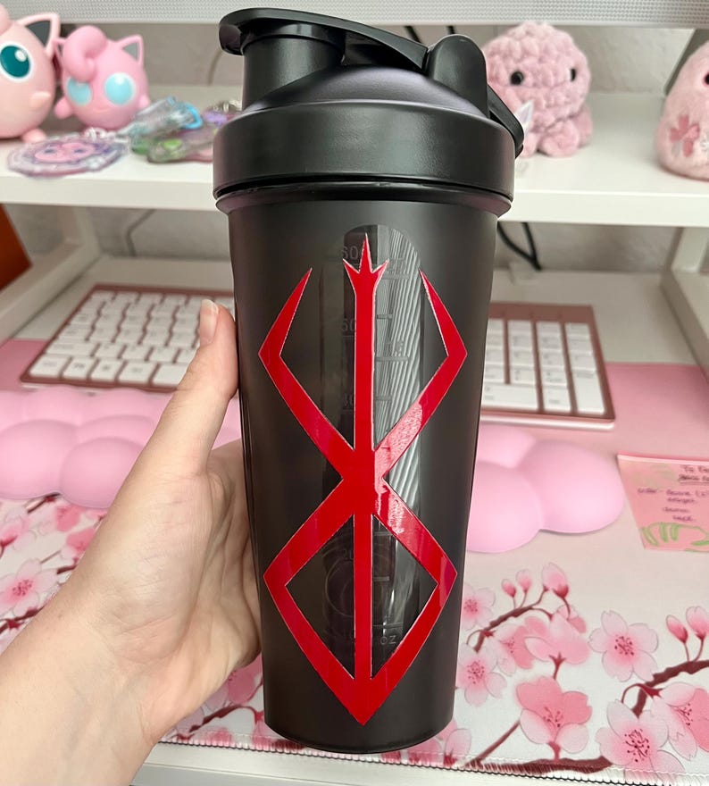 May include: Black shaker bottle with a red geometric design. The bottle has a black lid and a clear measurement scale on the side. The design is a red, stylized symbol. The bottle is held in a hand.