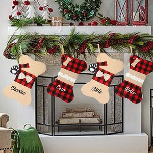May include: Four personalized dog bone-shaped Christmas stockings with red and black buffalo plaid fabric and beige fabric. The stockings are hung on a fireplace mantel with a garland of greenery and red berries. The stockings are personalized with the names "Charlie", "Coco", "Milo", and "Bella".