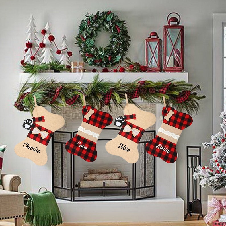 May include: Four personalized dog bone-shaped Christmas stockings with red and black buffalo plaid fabric and beige fabric. The stockings are hung on a fireplace mantel with a garland of greenery and red berries. The stockings are personalized with the names "Charlie", "Coco", "Milo", and "Bella".