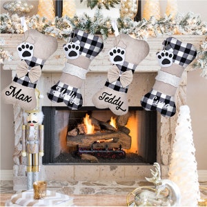 May include: Four personalized dog bone-shaped Christmas stockings with black and white buffalo plaid fabric and a bow. The stockings are hung on a fireplace mantel and are labeled "Max", "Molly", "Teddy", and "Luna".