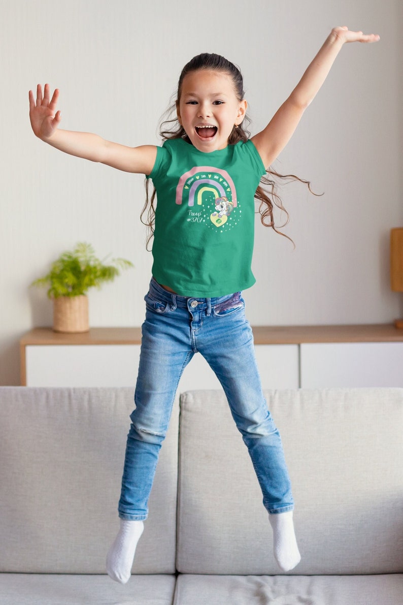 May include: A green t-shirt with a rainbow and unicorn graphic. The shirt has the text "Troop 3717". The child is wearing blue jeans and white socks. The child is jumping with arms raised.