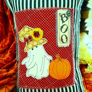 Puede incluir: Un cojín decorativo rectangular con un borde de rayas blancas y negras y un panel central de tela acolchada roja. El panel muestra un fantasma blanco con un sombrero amarillo estampado con una flor, una calabaza naranja y una etiqueta vertical que dice BOO.