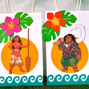May include: Two colorful gift bags with images of Moana and Maui from the Disney movie Moana. The bags feature a tropical theme with green leaves, flowers, and ocean waves.