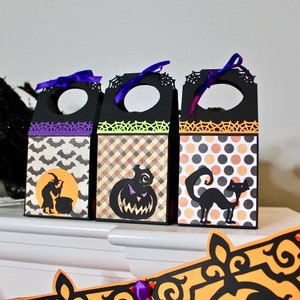 Puede incluir: Tres cajas de regalo negras de Halloween con recortes decorativos y detalles morados, verdes y naranjas. Cada caja presenta un diseño diferente: murciélagos, una calabaza y un gato negro. Un cuervo negro está sentado a la izquierda.