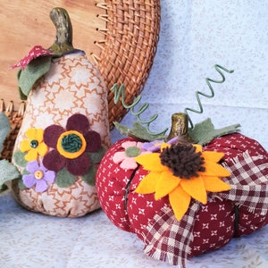 Puede incluir: Dos calabazas de tela con detalles de flores de fieltro. Una es de color beige con un estampado de estrellas y flores burdeos. La otra es roja con un estampado de estrellas blancas y un girasol amarillo. Ambas tienen tallos y hojas verdes.