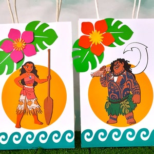May include: Two colorful gift bags with images of Moana and Maui from the Disney movie Moana. The bags feature a tropical theme with green leaves, flowers, and ocean waves.