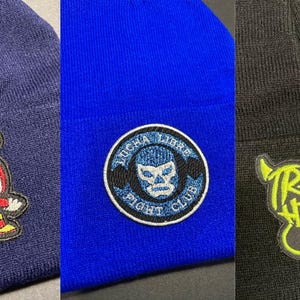 May include: Three different knit beanies with embroidered patches. The first beanie is navy blue with a red and yellow cartoon character. The second beanie is royal blue with a black and white Lucha Libre Fight Club patch. The third beanie is black with a yellow and black Trakas HDSPM patch.